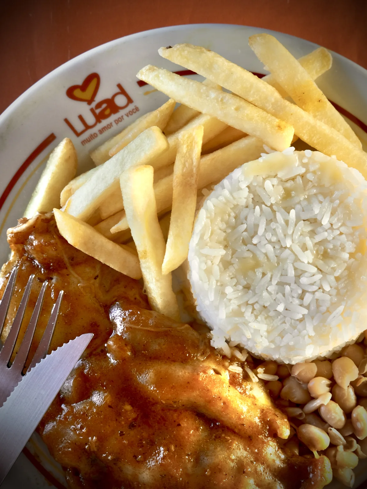 Bife à parmegiana com arroz, feijão e batata frita, servido no prato da Luad com o logo impresso